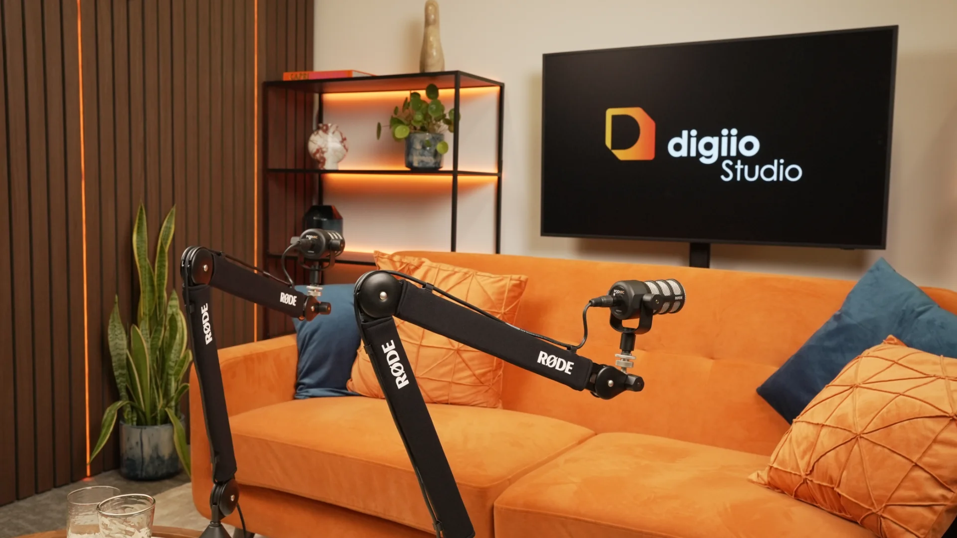 Two boom-arm microphones in front of the orange sofa, the digiio Studio logo on the screen behind