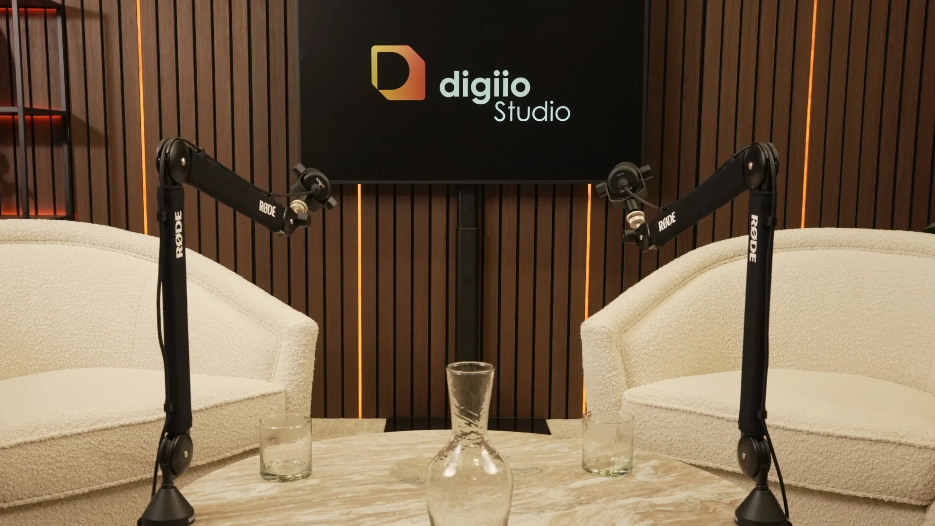 Two white armchairs facing across a marble table, microphones set, the studio logo on the screen between them